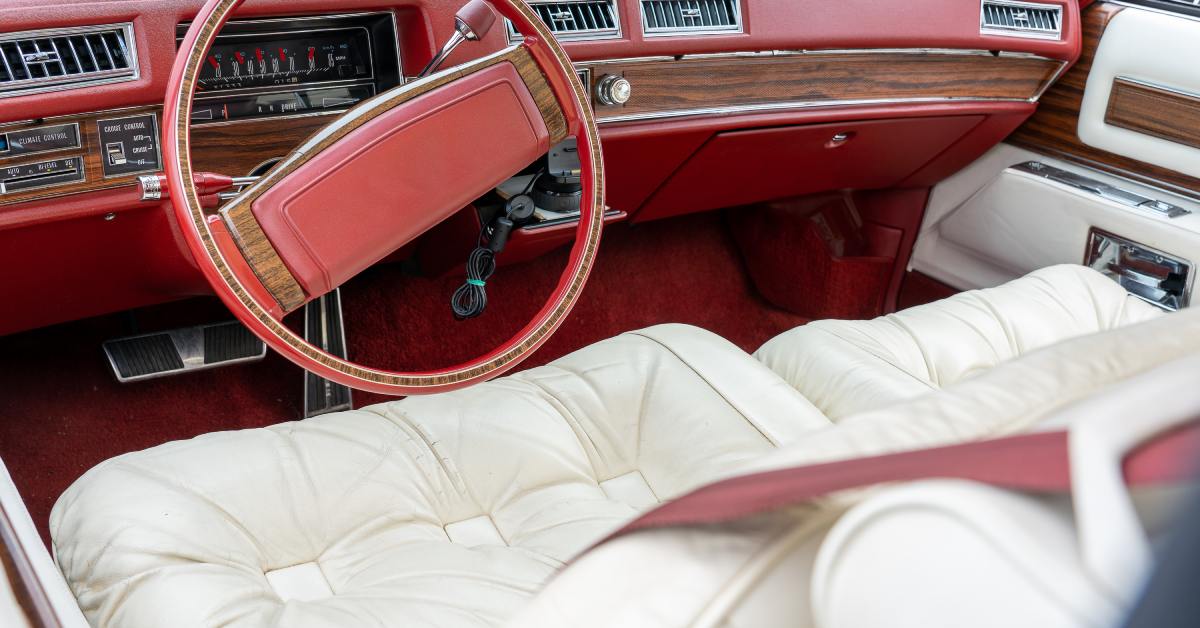 Interior of a vintage car with a red dashboard, wood trim, a large steering wheel, and white leather bench seats.