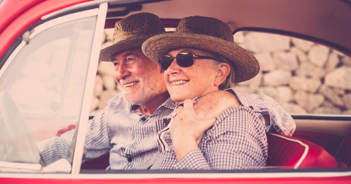 A smiling older adult couple in matching checkered shirts and straw hats sitting inside a classic red vehicle.