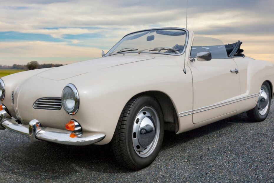 A white, vintage convertible Karmann Ghia with chrome bumpers parked on asphalt next to a green field.