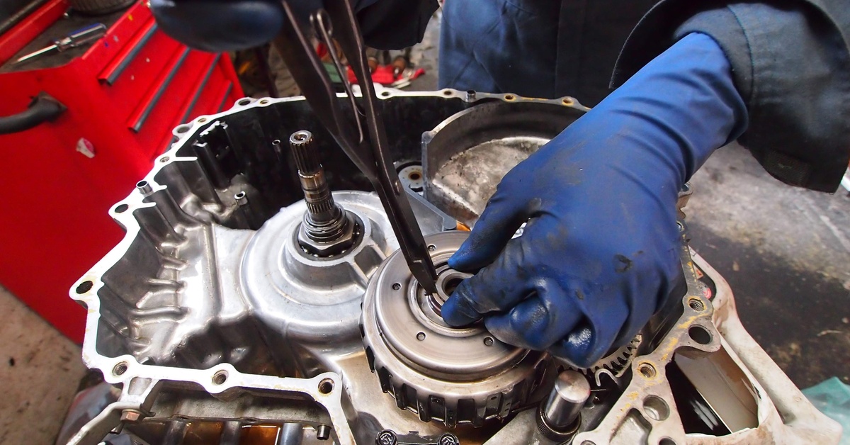 A close-up on a person's blue-gloved hands working on transmission parts of a car with a toolbox in the background.