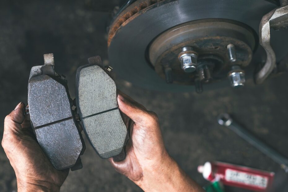 A person holding a brake pad in each hand, comparing the two. A wheel and various car tools rest in the background.