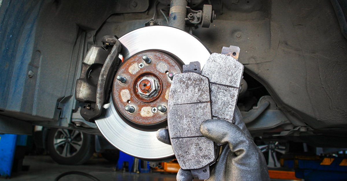 A hand holding two car brake pads in front of a vehicle's dusty wheel well with the tire removed.
