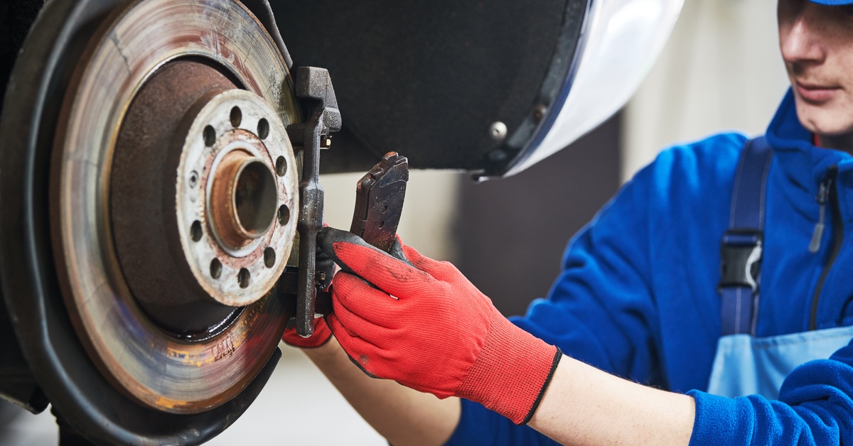 A man wearing a blue jacket and overalls replaces the brake pads around a car's wheel with various parts removed.