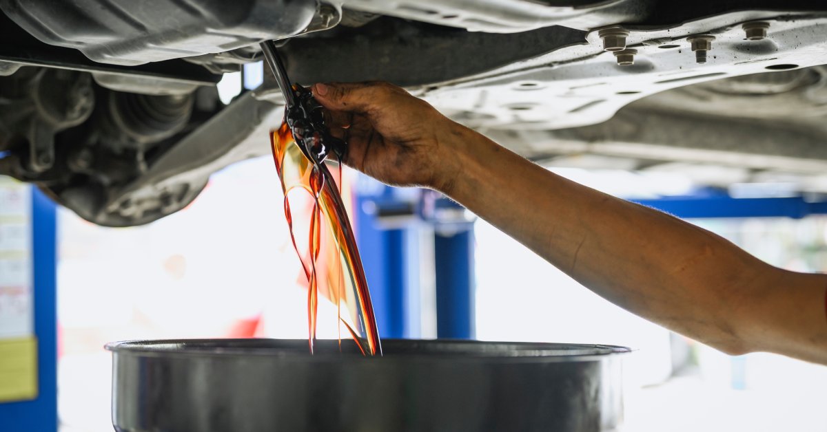 A close-up of a hand removing a part from underside a vehicle, draining the dirty engine oil into a collection pan.