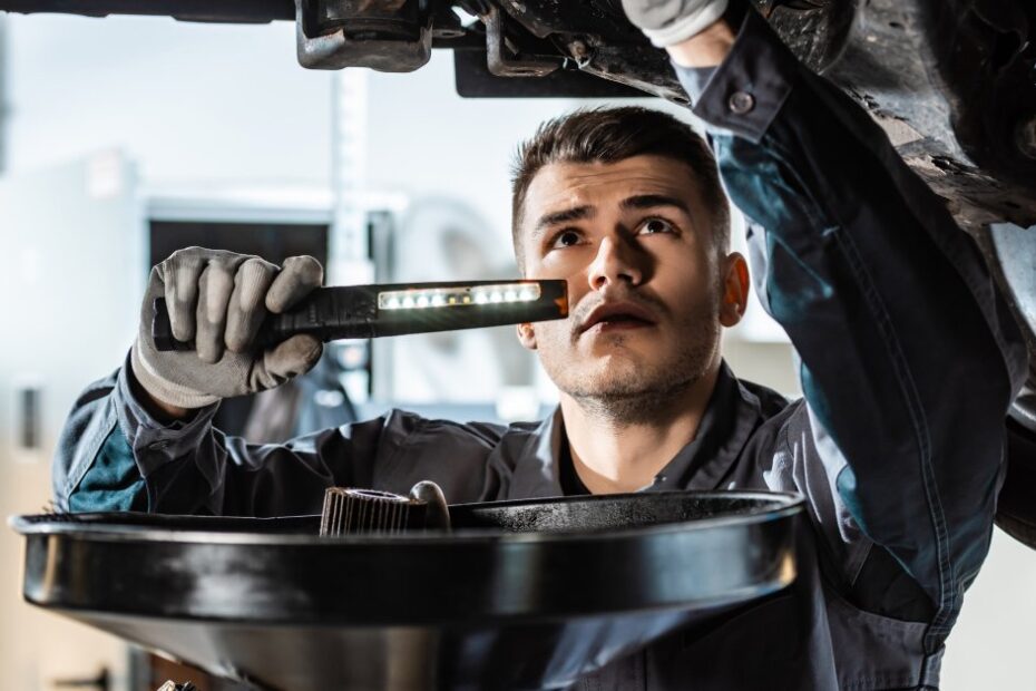 A young man with a handheld flashlight inspects the underside of a suspended vehicle, oil pan in front of him.
