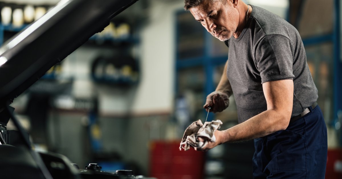 A man leans over the open hood of a vehicle, dip stick in hand, examining the oil from the vehicle.