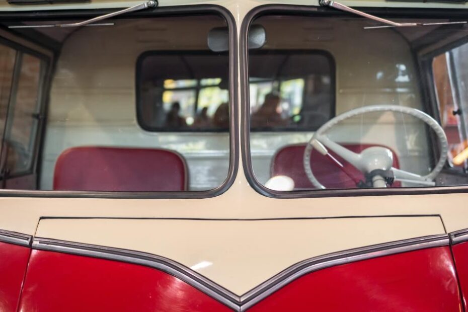 A close-up of the windshield of a classic bus, looking in at a red interior seating and a tan steering wheel.