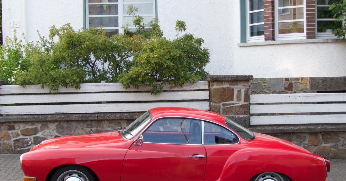 A red Karmann Ghia Coupe sits parked outside on the road next to a white and brick apartment building.