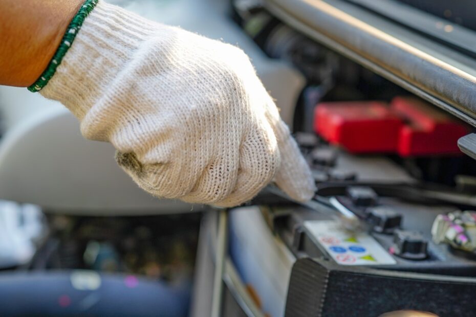 A close-up of a gloved hand inspecting a car's battery with a tool under the open hood of the vehicle.