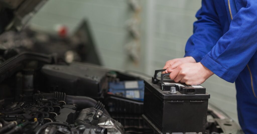 A person in a blue jumpsuit lifting a battery out of a car with an open hood, showing the engine components.