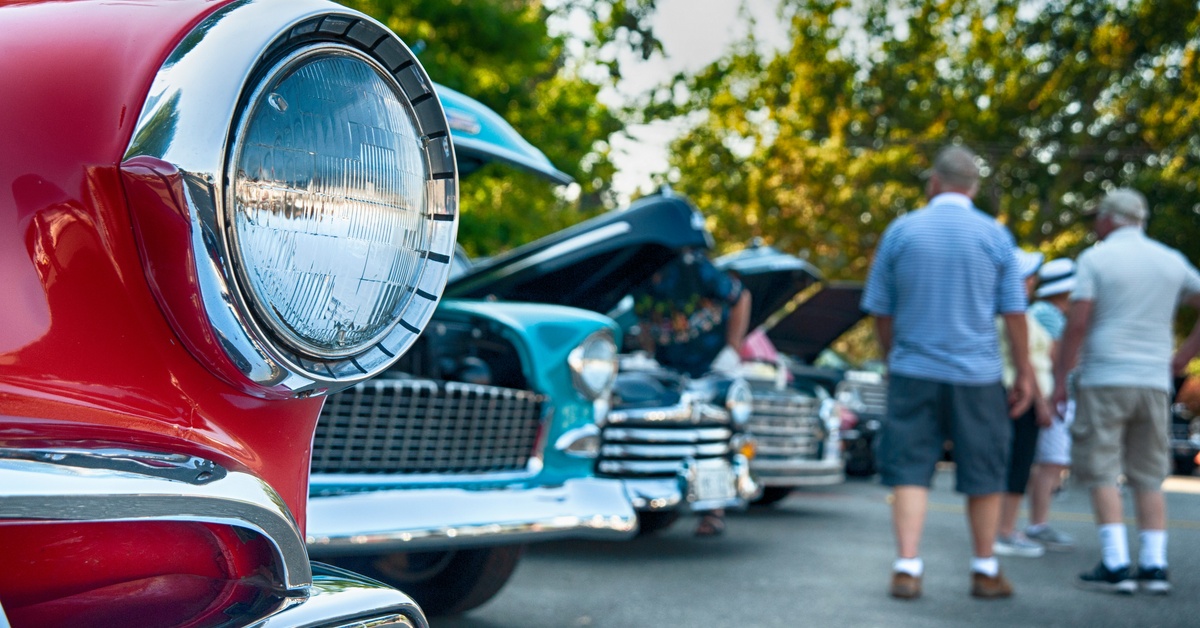 A close-up of a car's headlight with several other classic cars lined up behind it and people walking the lot.