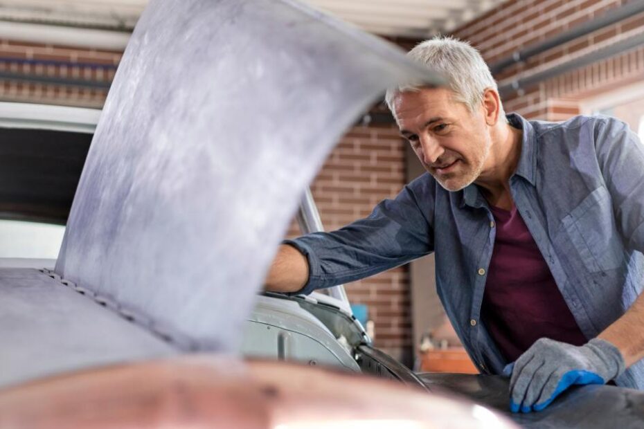 A man with graying hair wearing gloves opens the hood of a classic car in a brick garage building.
