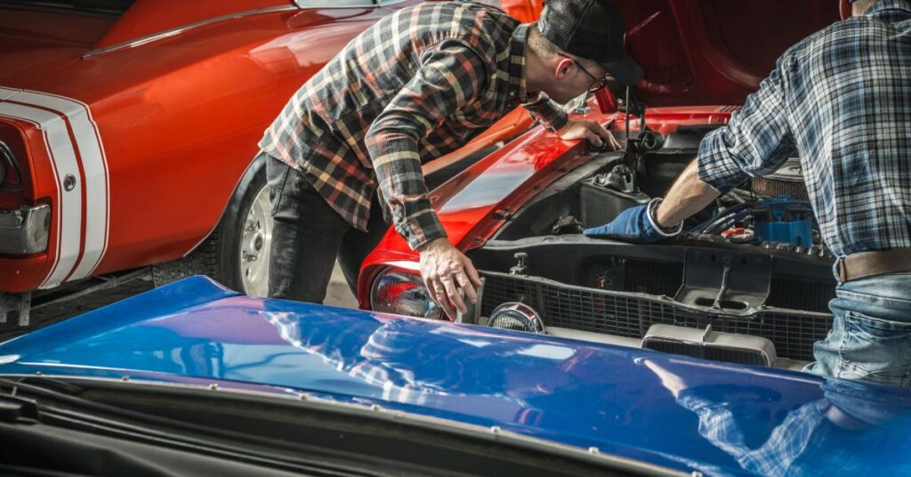 Two men wearing plaid shirts working under an open hood of a classic car in a shop with other cars surrounding them.