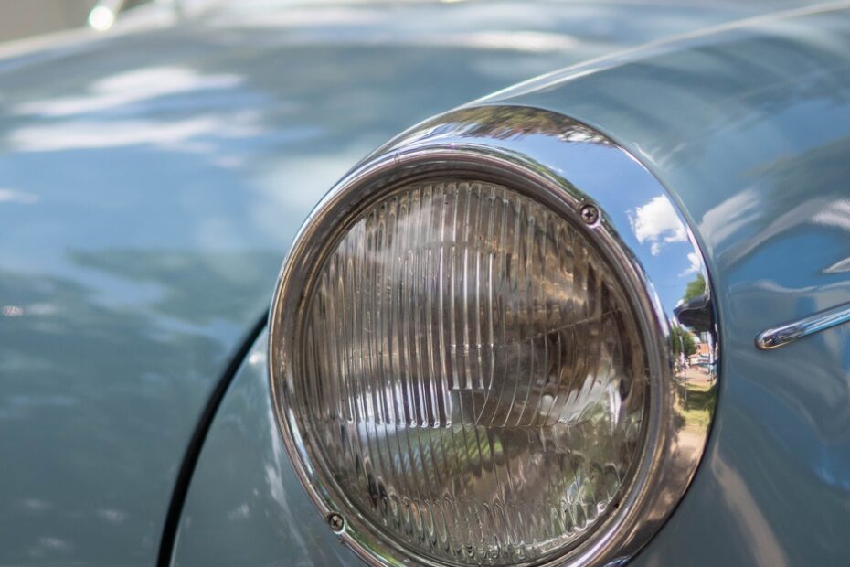 A close-up of a round headlight on a classic, slate blue car. The paint is shiny and reflects the sunlight.