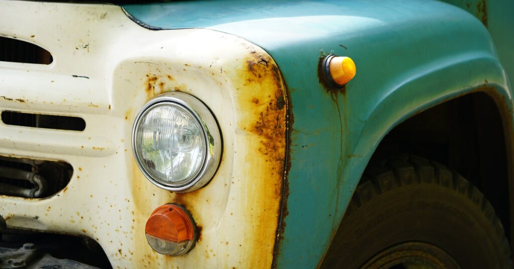 A close-up of a rusty front fender of an old car with white and blue paint. A headlight and part of the grille are also rusted.