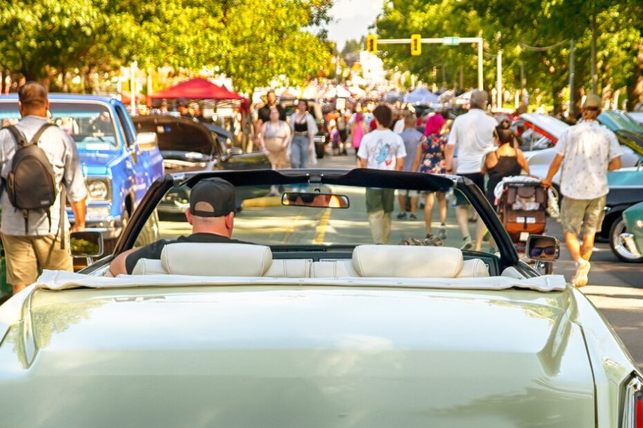 A man wearing a ball cap drives a classic car down a crowded road with cars parked along either side at a classic car show.