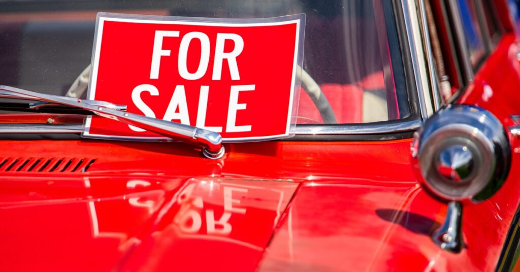 A red car's hood and lower right corner of the windshield with a "FOR SALE" sign pinned by the classic windshield wiper.