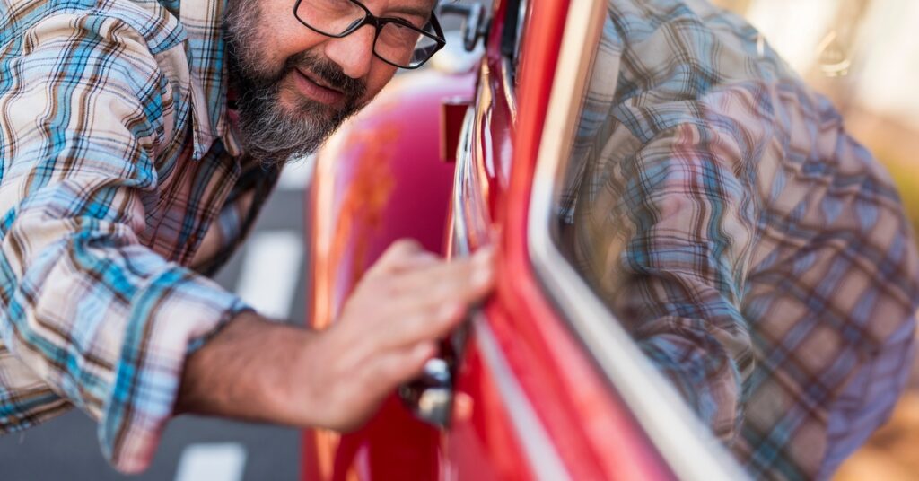 A close-up of a man with glasses in a plaid shirt inspecting the door of a classic, red car in the outdoor sun.