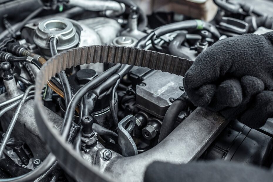 A close-up of two gloved hands holding a replacement belt over the inner components of a car's under-hood space.