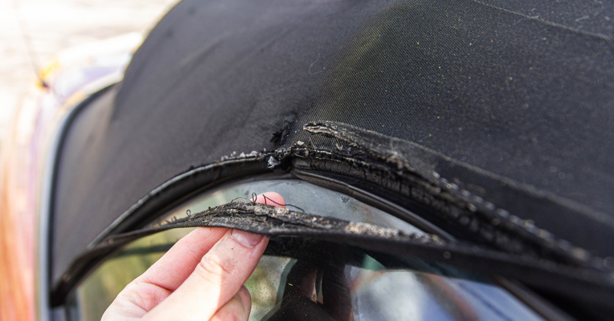 A close-up of a hand holding the edge of a black fabric convertible top that has frayed and is peeling away from the rest.
