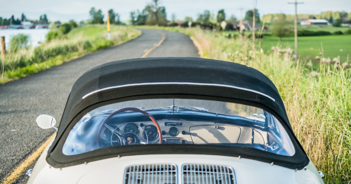 The rear window of a classic car with a black convertible fabric top parked on the side of the road near tall grass.