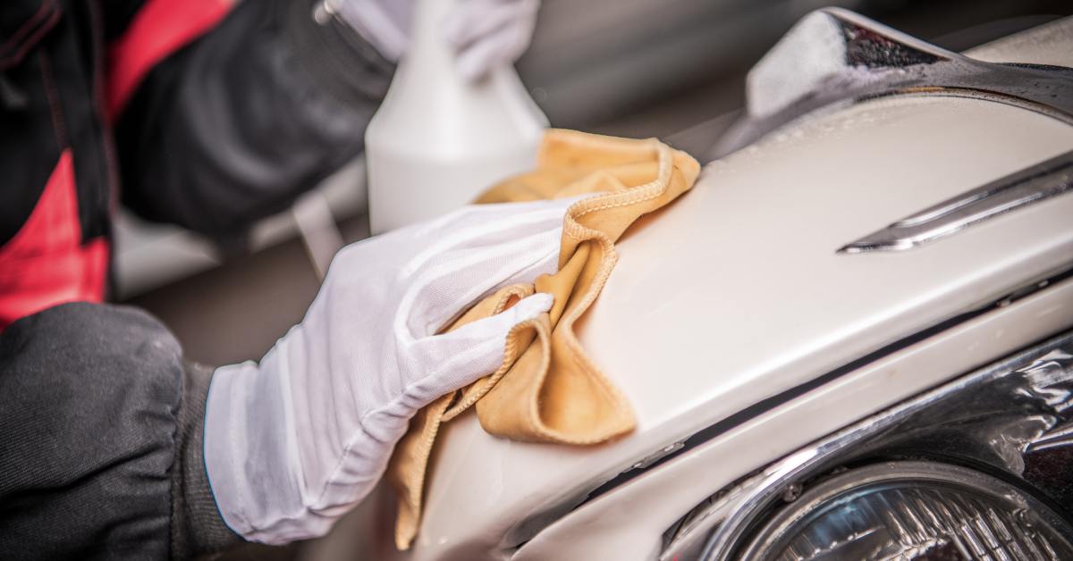 A person holding a spray bottle and wearing white silk gloves while cleaning the exterior of a white vintage car.