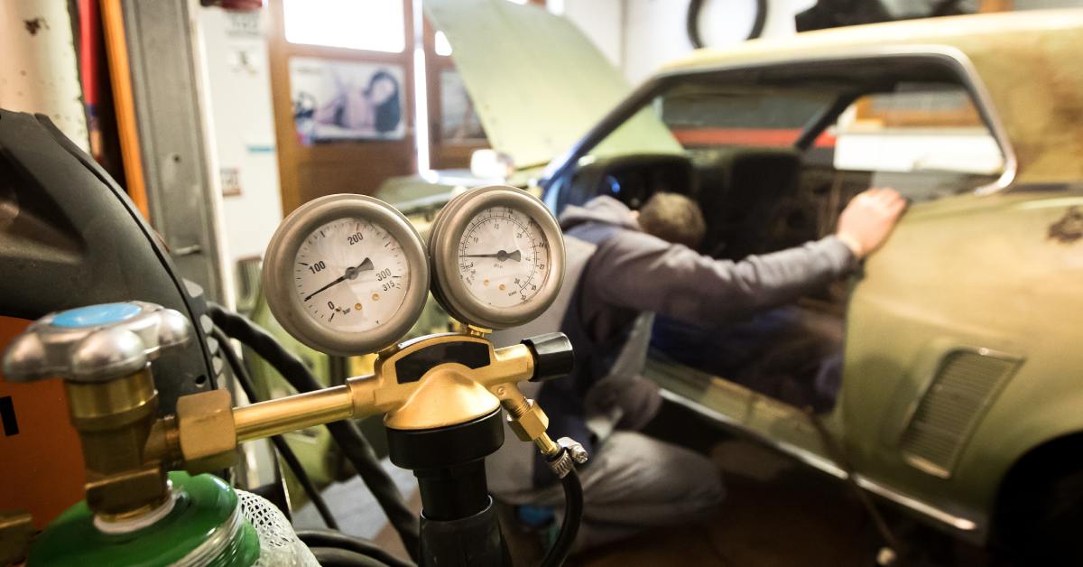 A man crouched down to inspect the interior of a classic car in a workshop. A pressure tank sits in the foreground.