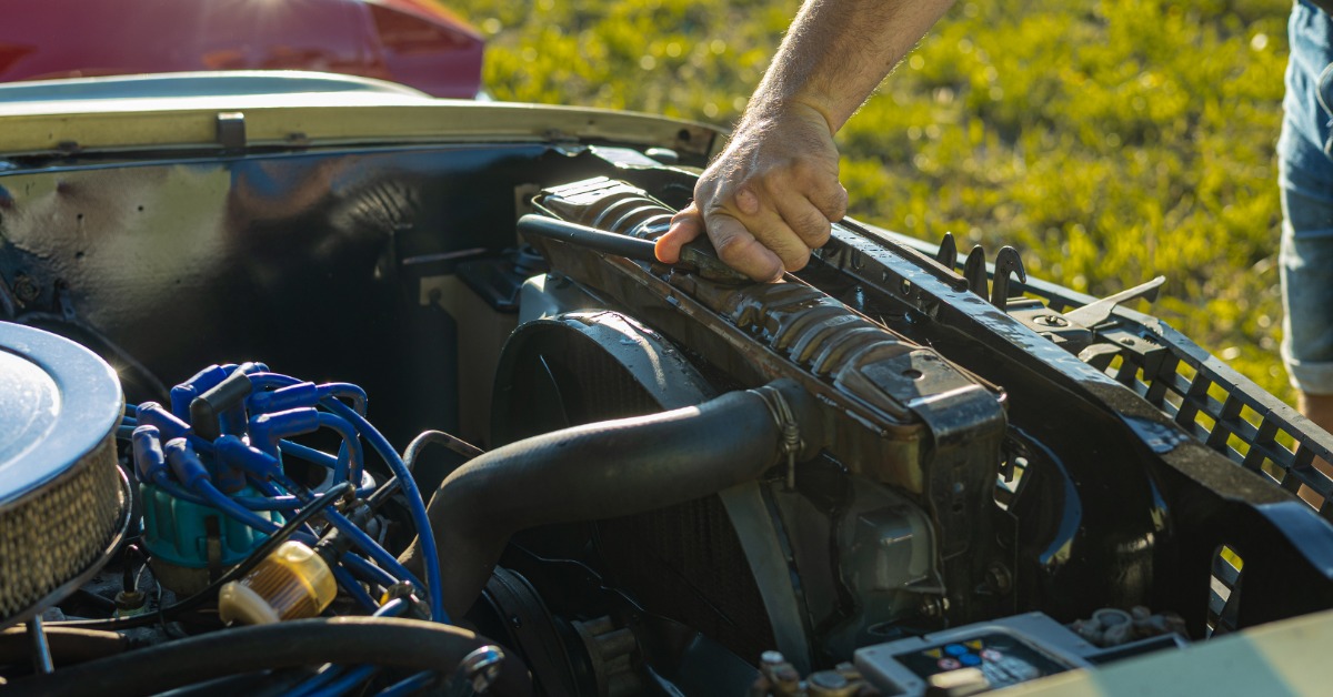 A man's arm untwists a cap on the open engine compartment of a vintage car against a blurry, grassy background.