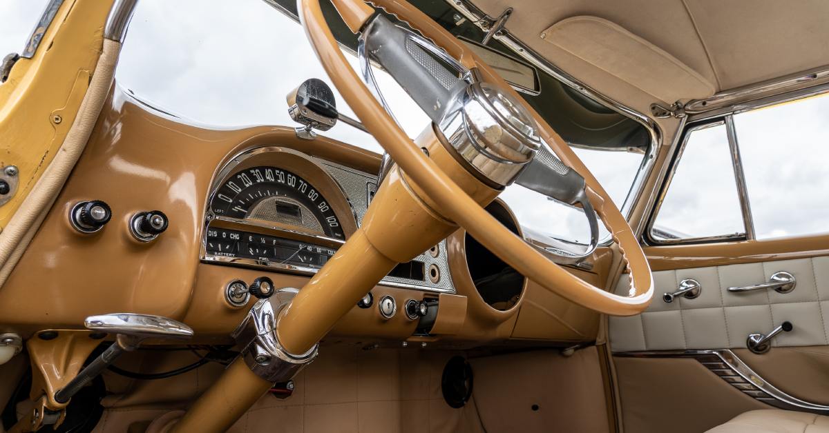 A close-up of the steering wheel in the interior of a classic car, colored in beiges and light brown colors.