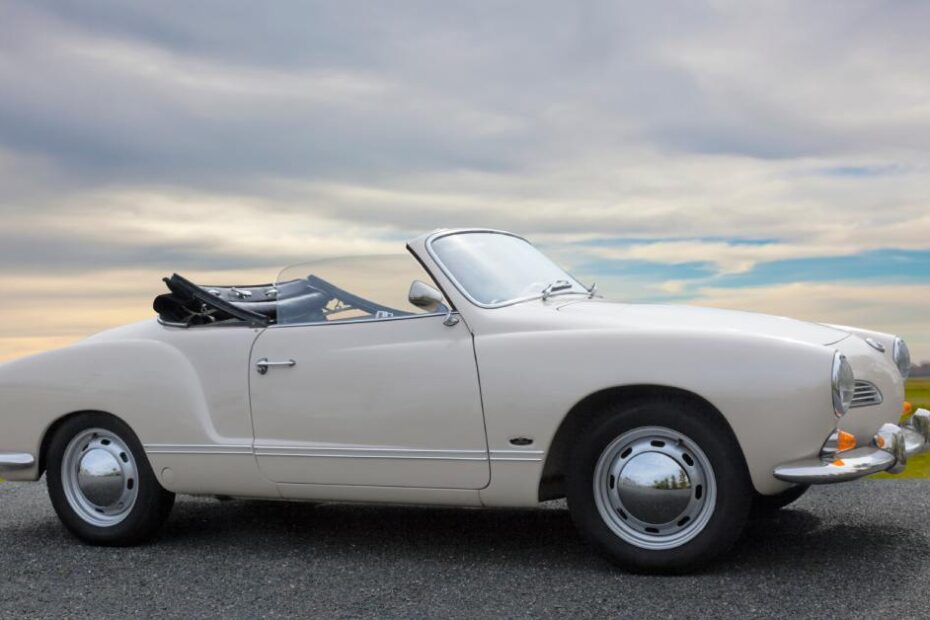 A VW Karmann Ghia convertible model car with an off-white paint job on a road near a grassy field with overhead clouds.