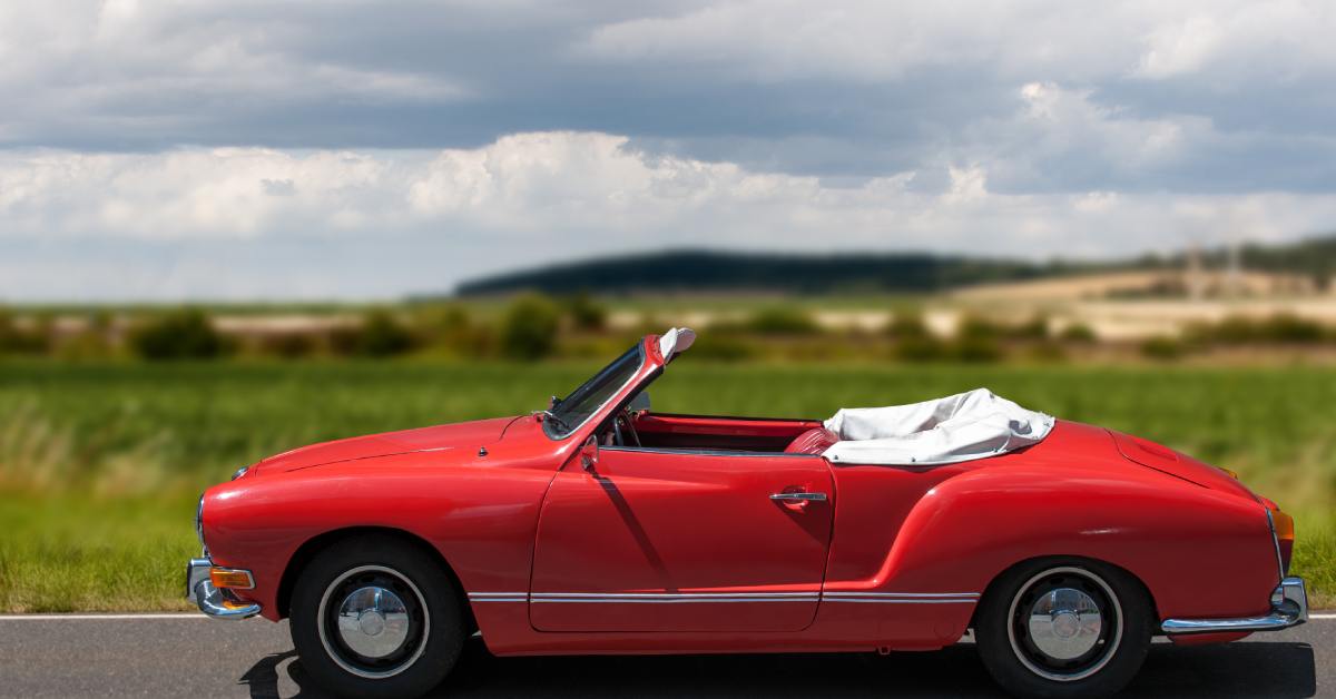 A Karmann Ghia Cabrio in a red paint color with a convertible roof, driving along a grassy landscape.